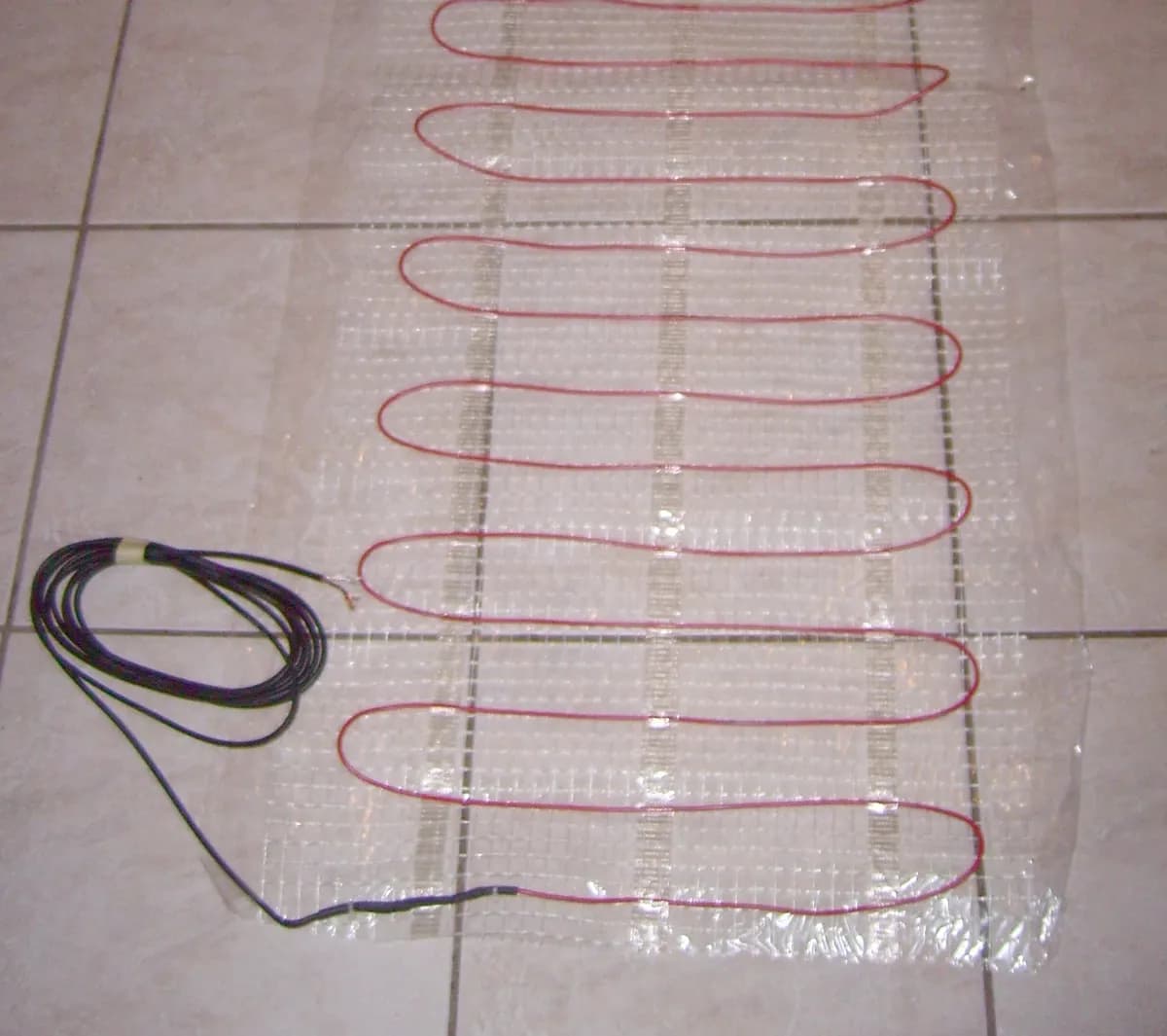 An electric floor heating mat unrolled on a tile floor: a red heating cable woven back and forth across clear mesh, with the black power lead coiled at one corner