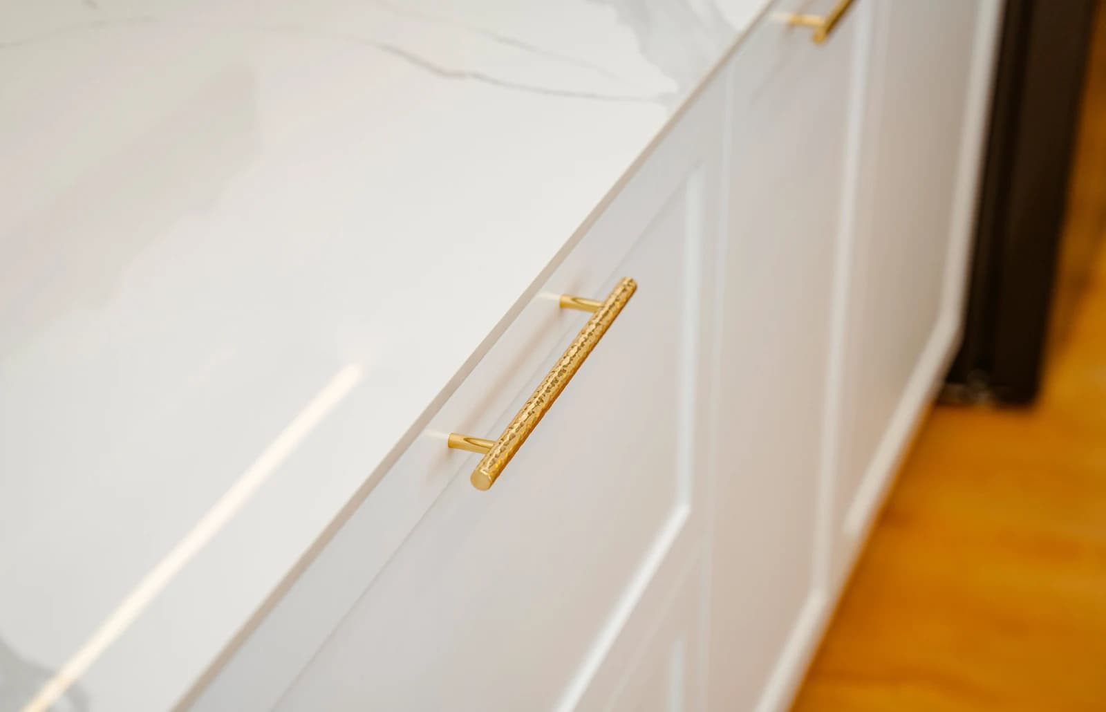 Close-up of white Shaker-style cabinet drawer fronts with a hammered brass bar pull, under a white marble-look countertop with grey veining