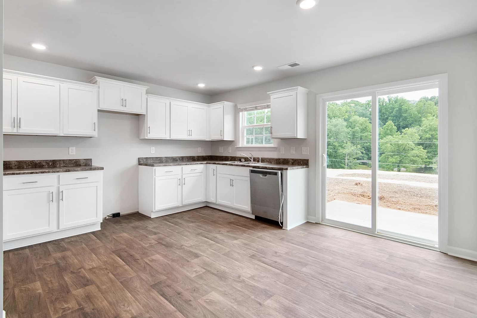 Empty kitchen with white Shaker-style upper and base cabinets with brushed-nickel bar pulls in an L-shape, dark brown speckled countertops, a stainless steel dishwasher, a wood-look plank floor and a sliding glass door looking out onto trees