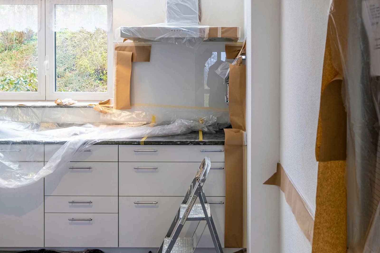 Kitchen in the middle of renovation, with white flat-front base cabinets in place under a dark countertop, the counter and walls covered in plastic sheeting and masking paper, and a folding step ladder in front