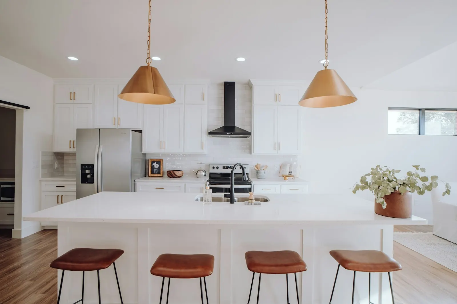 White Shaker-style kitchen island with an undermount sink and black faucet, a white countertop with a seating overhang, four leather bar stools, and two brass pendant lights