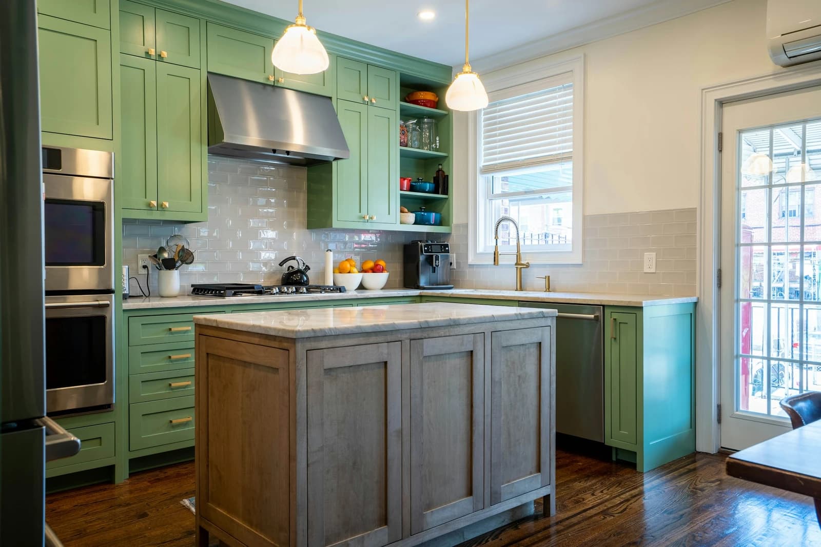 Kitchen with sage-green Shaker cabinets, a white subway tile backsplash, and a gray-washed wood island topped with a light veined natural stone countertop under two glass pendant lights