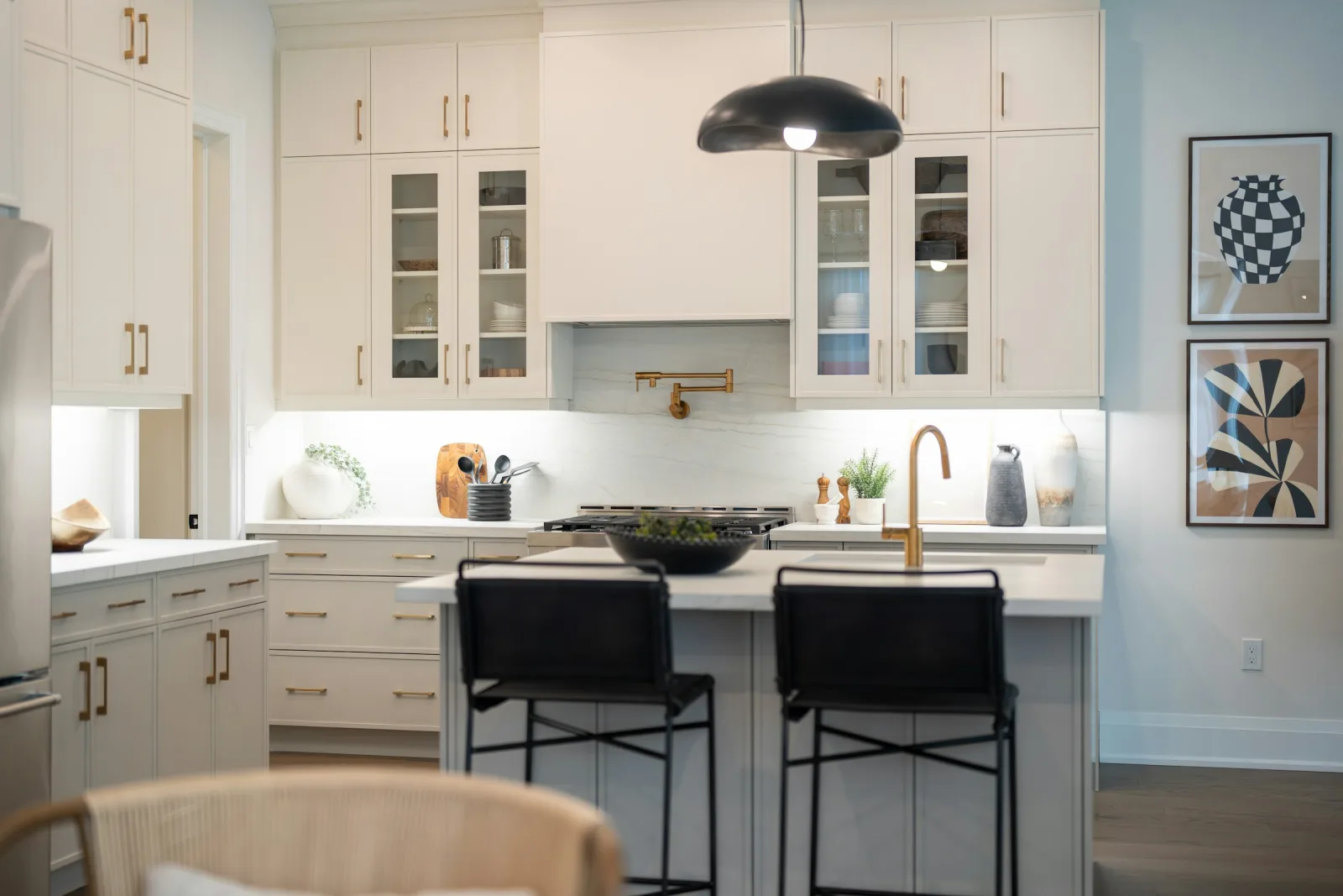 White Shaker-style kitchen with cabinets to the ceiling, glass-front uppers, a slab backsplash, a brass pot filler, and an island with two black bar stools