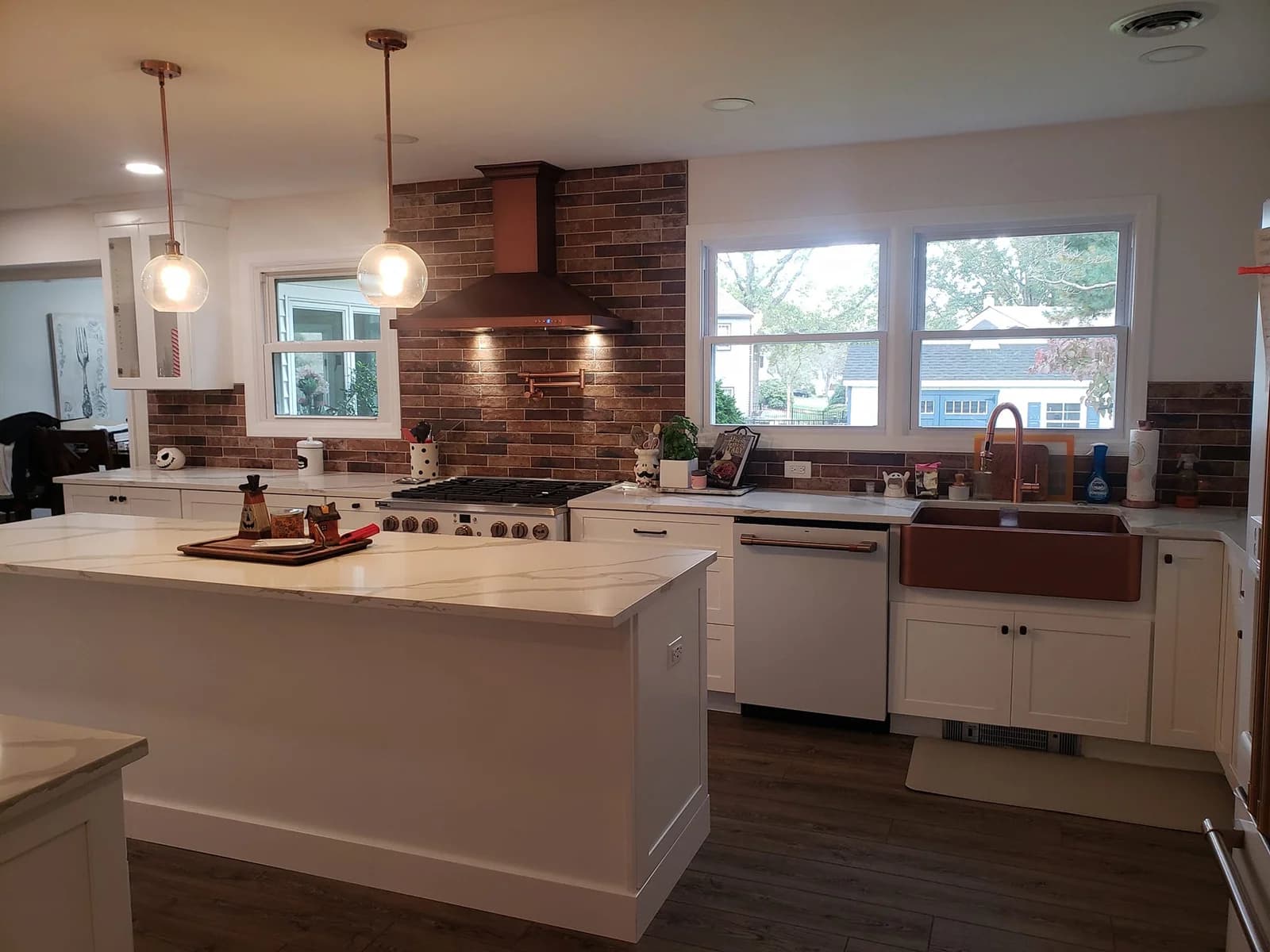 Foreverbuilt kitchen with a long white Shaker-style island, a white veined countertop, an outlet on the island's end panel, copper pendant lights, and a copper range hood