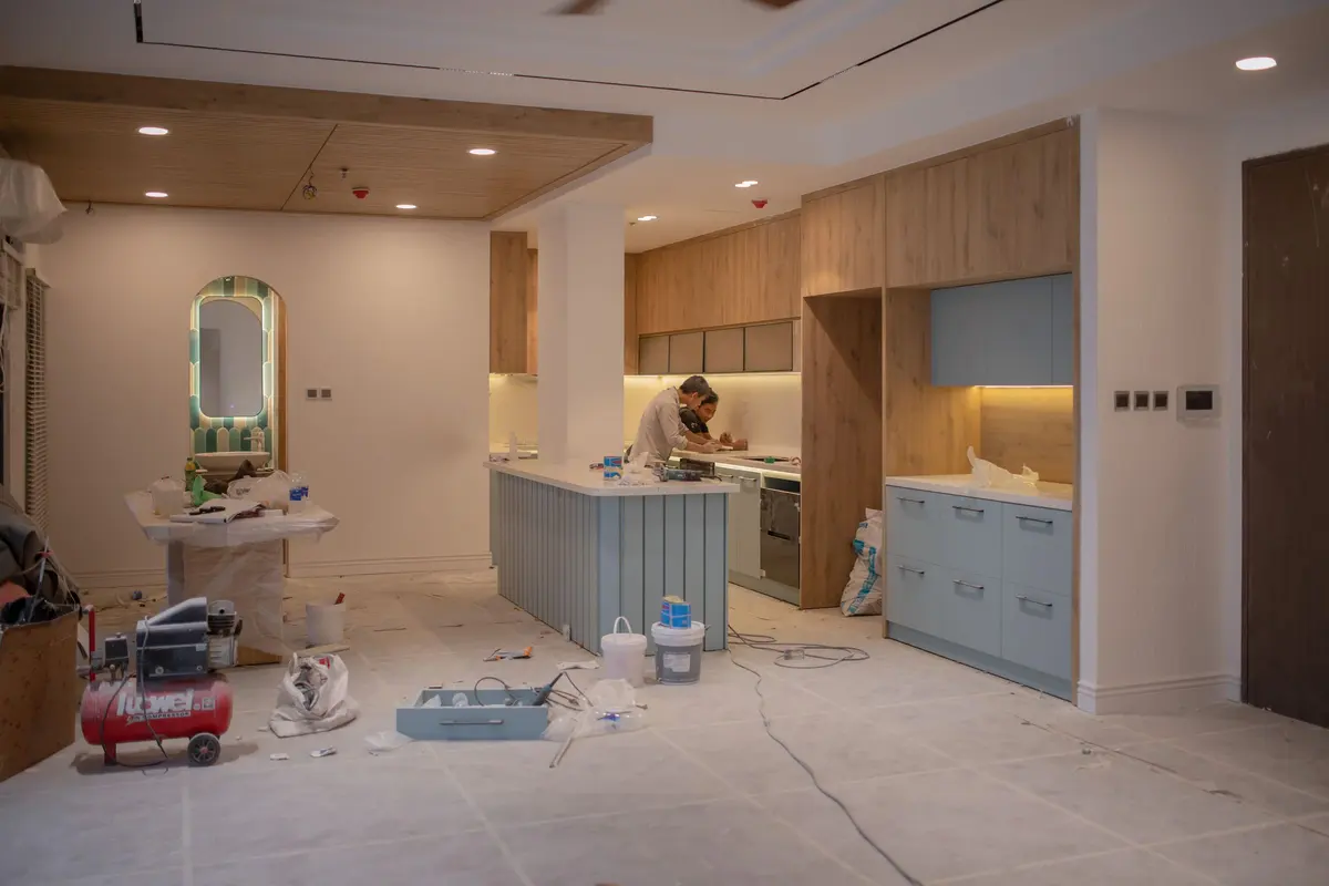 Kitchen under construction: two installers working at the counter behind a pale blue island, with tools, buckets, and an air compressor on the protected floor