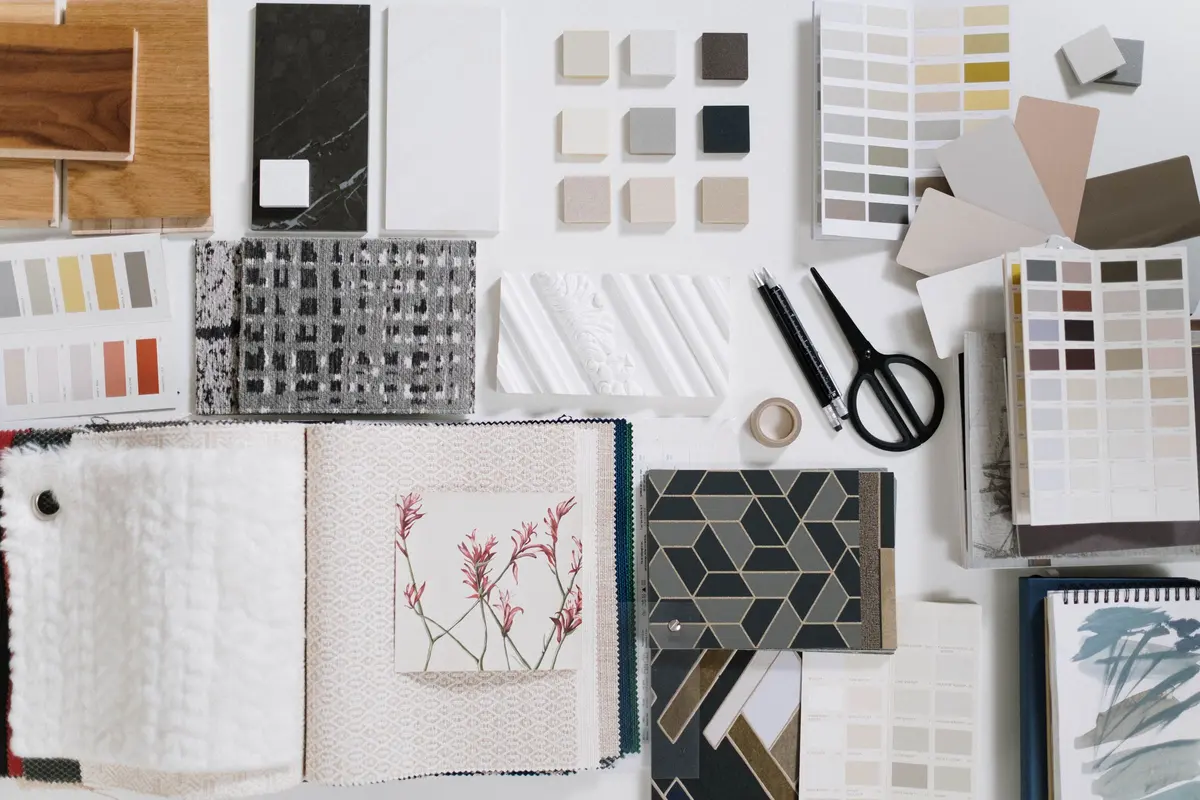 Flat lay of design samples on a table: tile chips, paint color cards, wood and stone swatches, fabric, and scissors