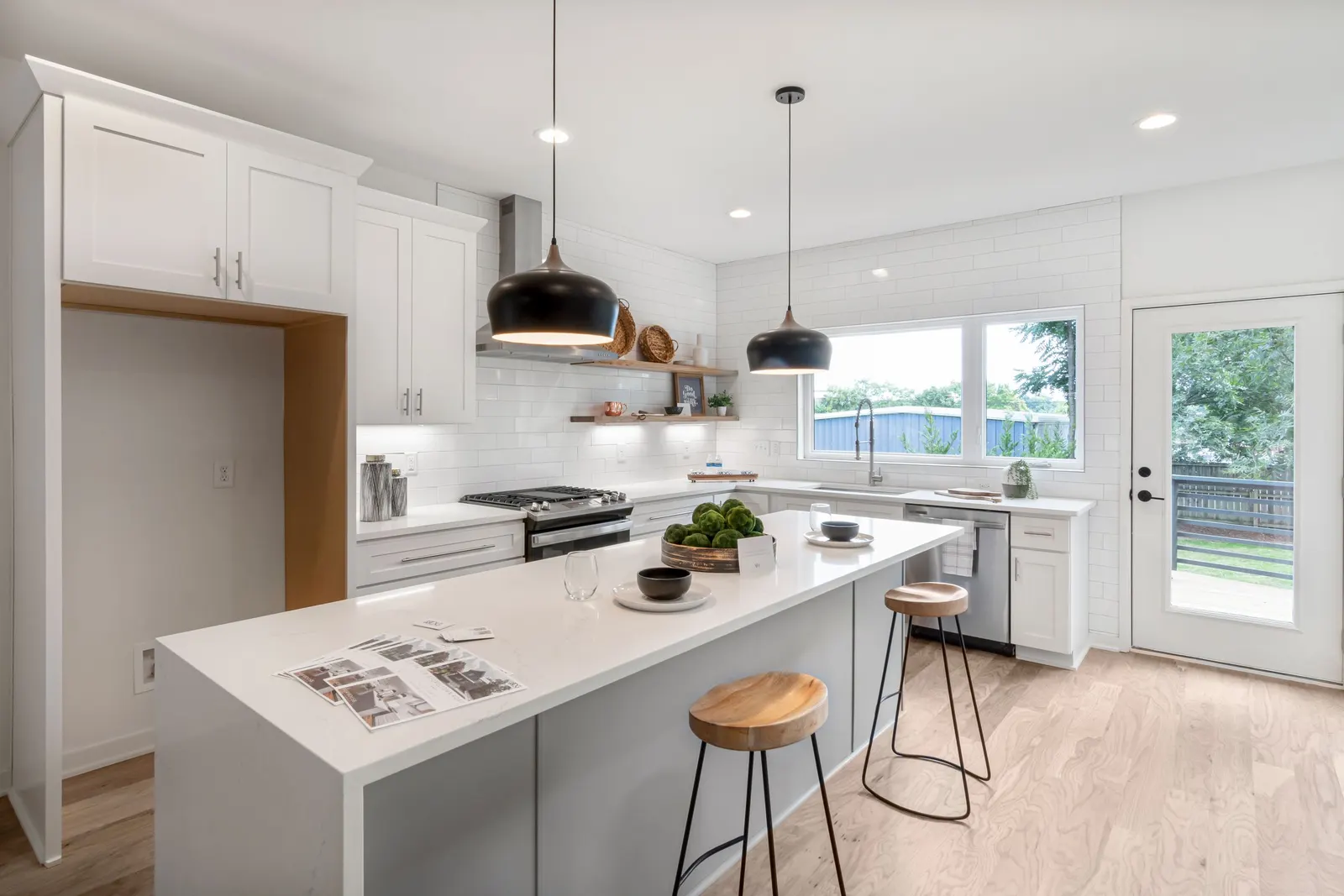 Bright kitchen with white Shaker cabinets, a white subway tile wall, a long white island with two wood stools, black pendant lights, and light oak floors